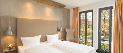 A hotel room with two beds, a large window with curtains, a bedside table with a lamp, and a view of the outside.