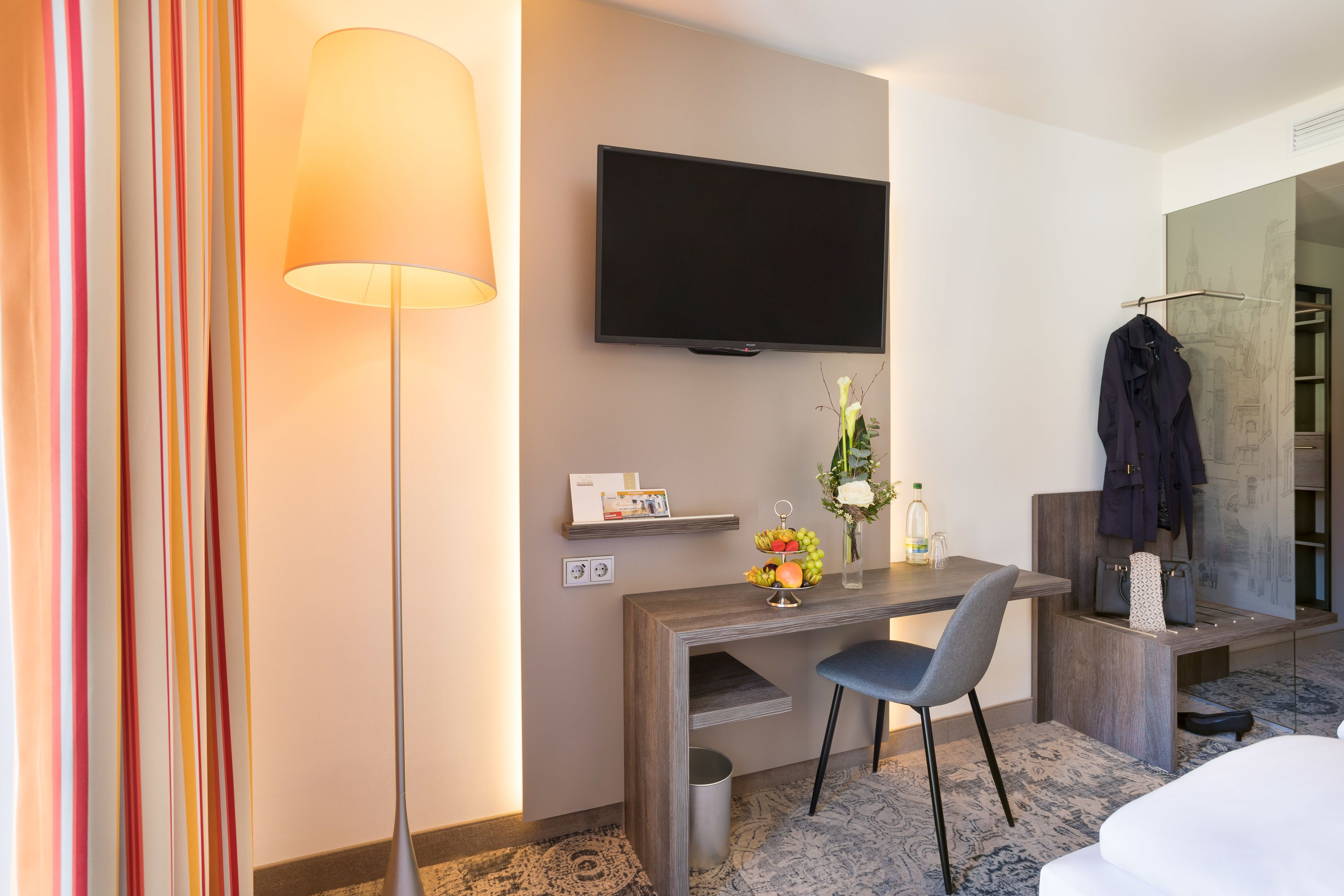 A hotel room with a desk, chair, TV, and a coat rack.