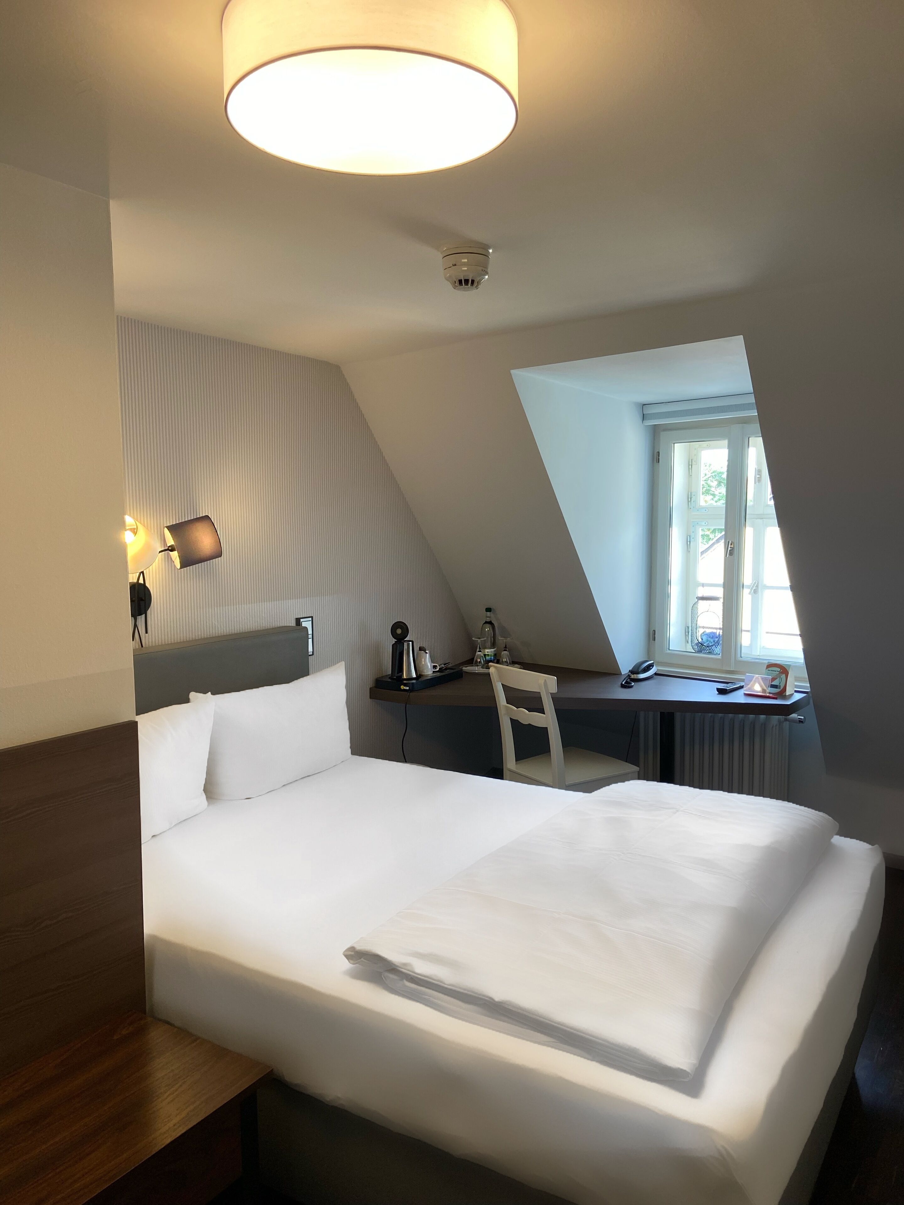 Ein Hotelzimmer mit einem ordentlich bezogenen Bett, einem Schreibtisch mit Stuhl, einer Lampe und einem an der Decke befestigten Rauchmelder.