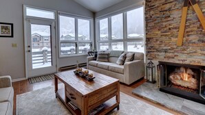 A cozy living room with a stone fireplace, a wooden coffee table, a beige sofa, and large windows offering a view of snow-covered houses. - Private Mountain Town home Pet Friendly with Great Views - WD12 by Redawning (Copper Mountain)