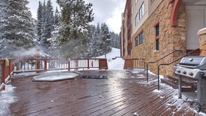 Una terraza de madera con un jacuzzi y una parrilla, rodeada de árboles cubiertos de nieve y un edificio de piedra.