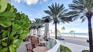 A seaside patio with tables and chairs, palm trees, and a view of the ocean. - 1201 W S 1 Bedroom Condo by RedAwning (Fort Lauderdale)
