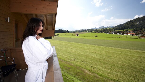A person in a white robe standing on a balcony with a view of a green field and houses in the distance. - Residenz Cristallo (Reit im Winkl)
