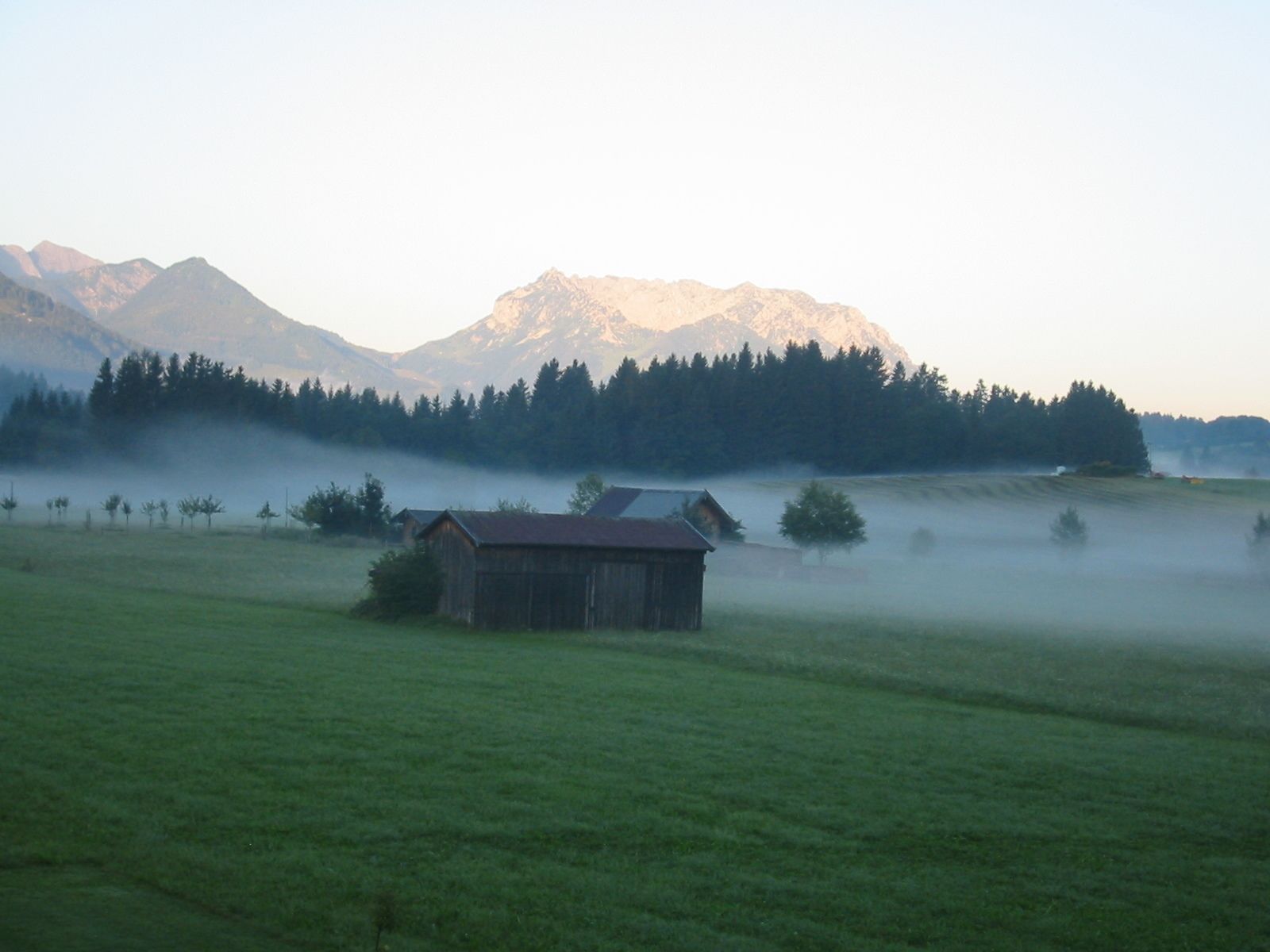 A rural landscape with a wooden barn in the foreground, a forested area, and mountains in the background.