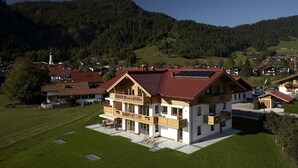A large, two-story chalet with a red roof, situated on a grassy hill with a forested mountain in the background. - Residenz Cristallo (Reit im Winkl)