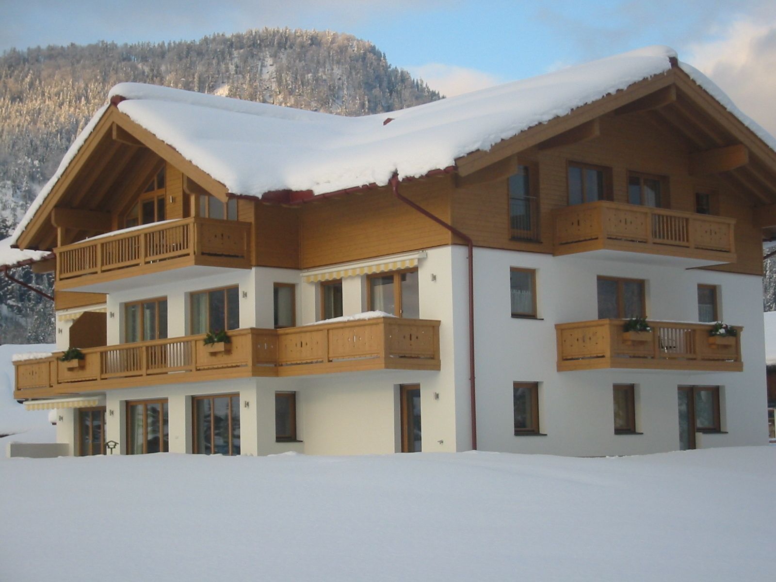 Ein mehrstöckiges Chalet mit Holzbalkonen und einem mit Schnee bedeckten Dach, vor einer Bergkulisse gelegen.
