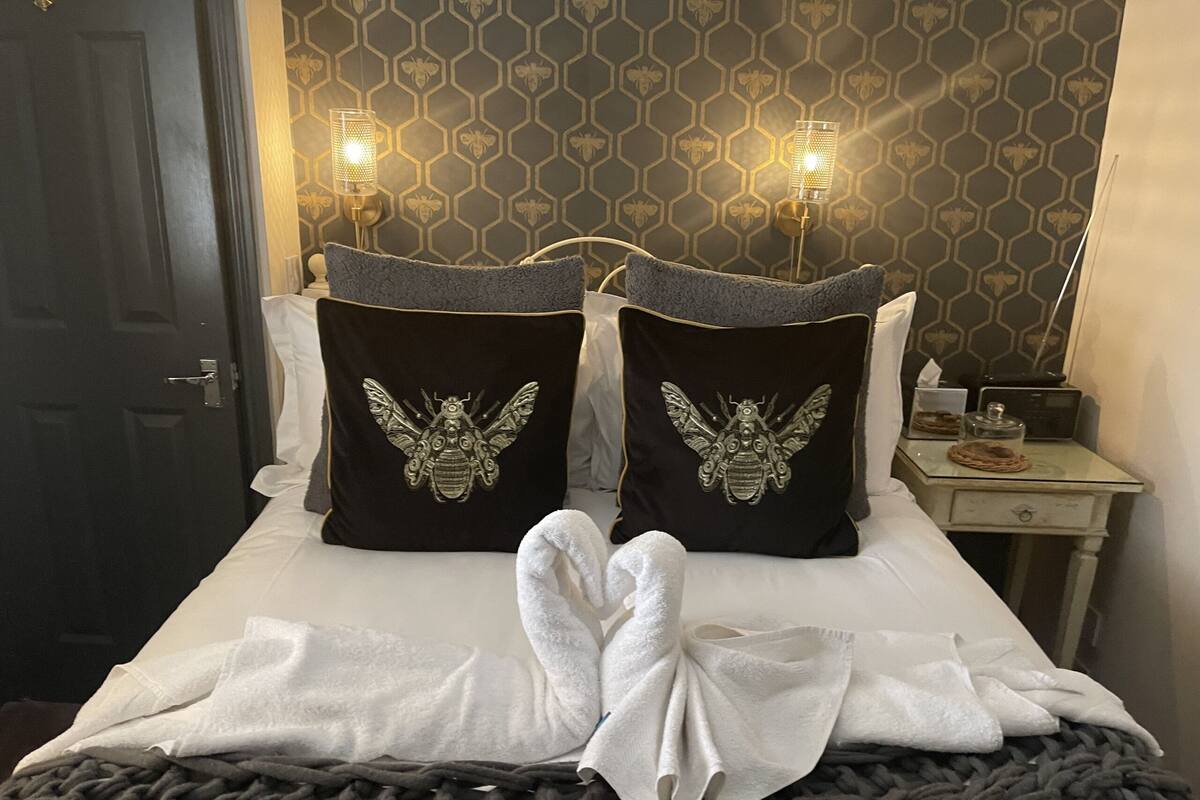 A neatly made bed with two decorative pillows featuring a bee design, a bedside table with a lamp, and a patterned wallpaper.