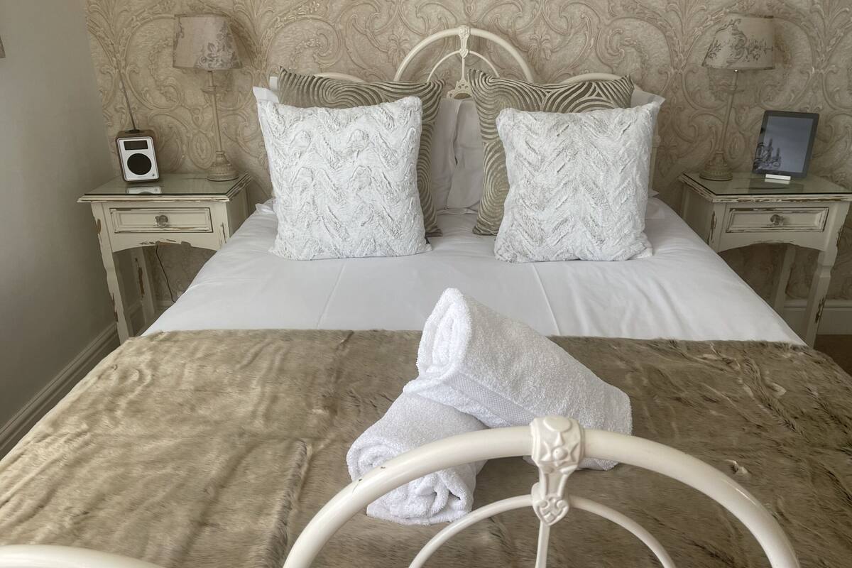 A neatly made bed with a white comforter and two patterned pillows, a white throw blanket, and a white towel on a metal bed frame.