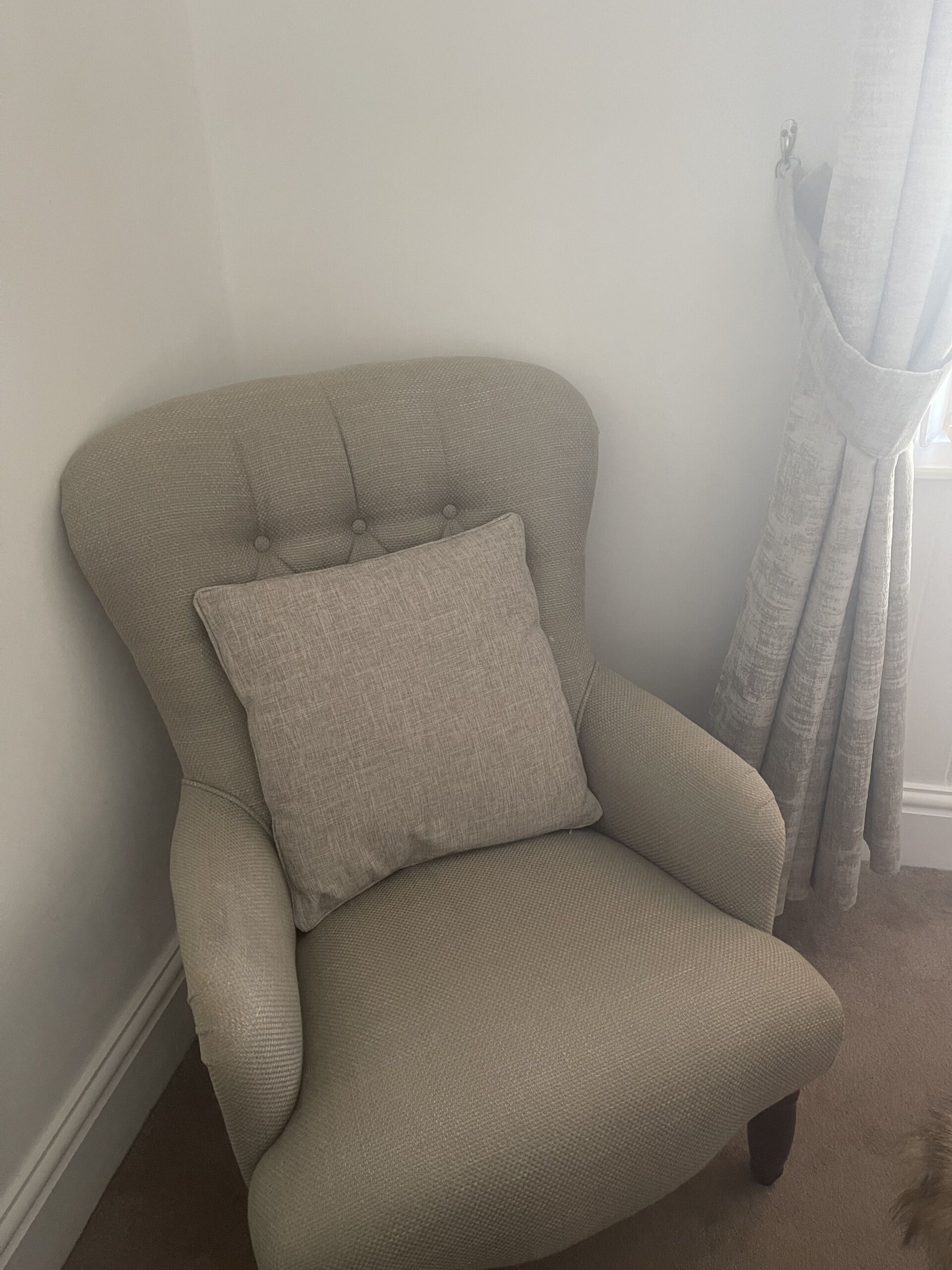 Un sillón beige con un cojín, colocado frente a una pared lisa con una ventana y una cortina.