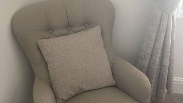 Un sillón beige con un cojín, colocado frente a una pared lisa con una ventana y una cortina.