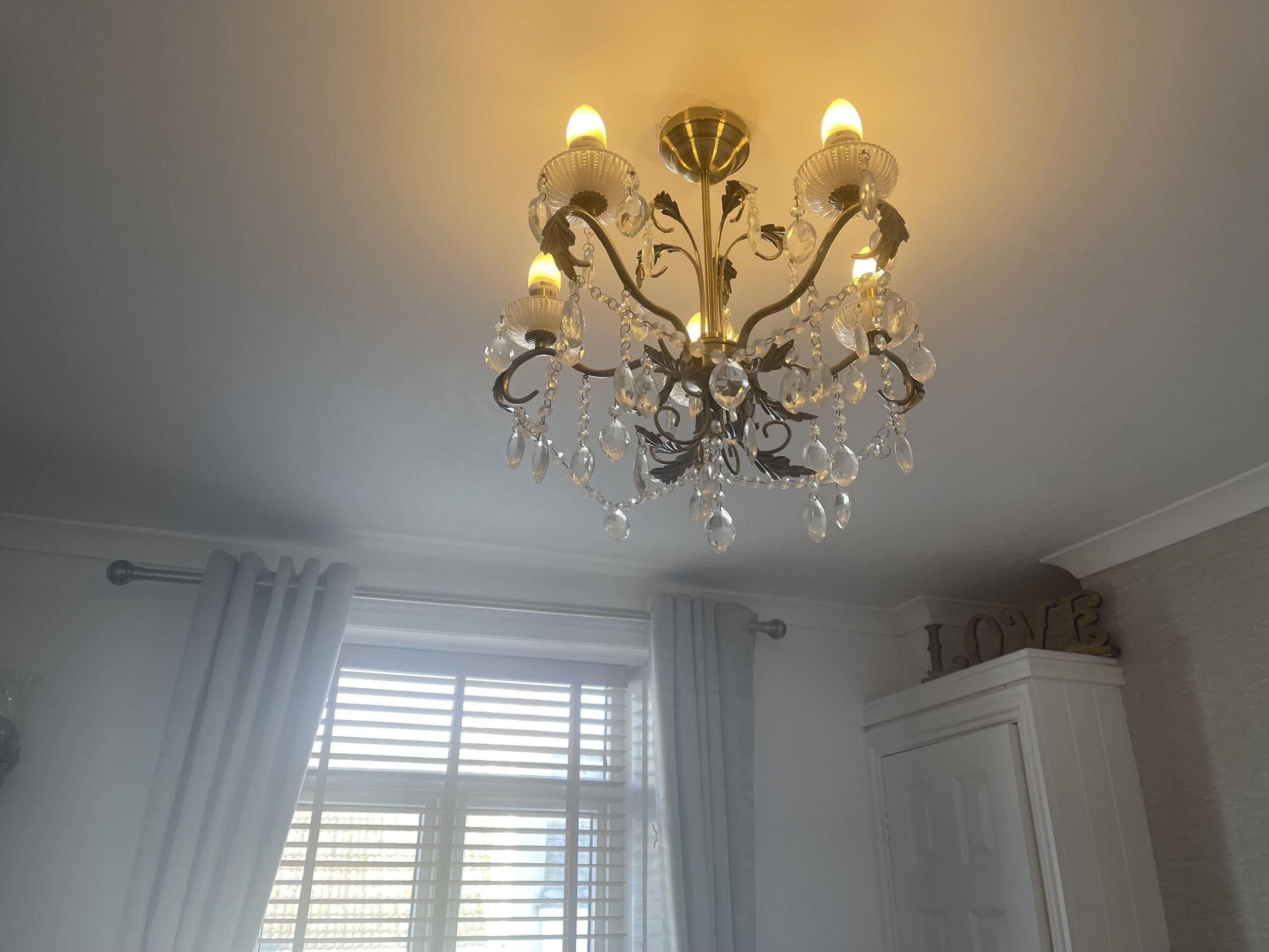 Un candelabro con múltiples brazos y detalles de cristal en una habitación con paredes blancas y una ventana con persianas.