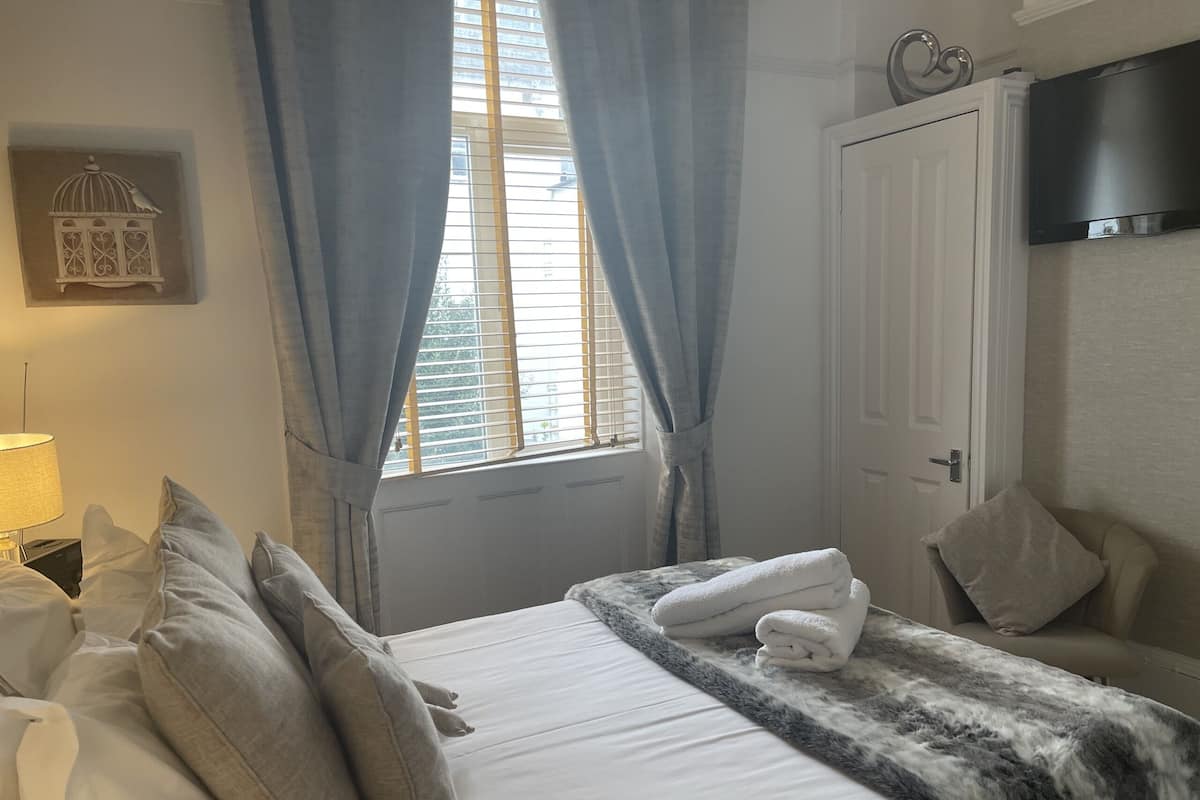 A neatly made bed with pillows, a bedside lamp, a window with curtains, and a television on the wall.