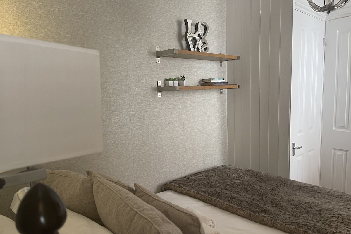 A bedroom with a bed, two pillows, a bedside table, a lamp, a wall-mounted shelf with decorative items, and a door.