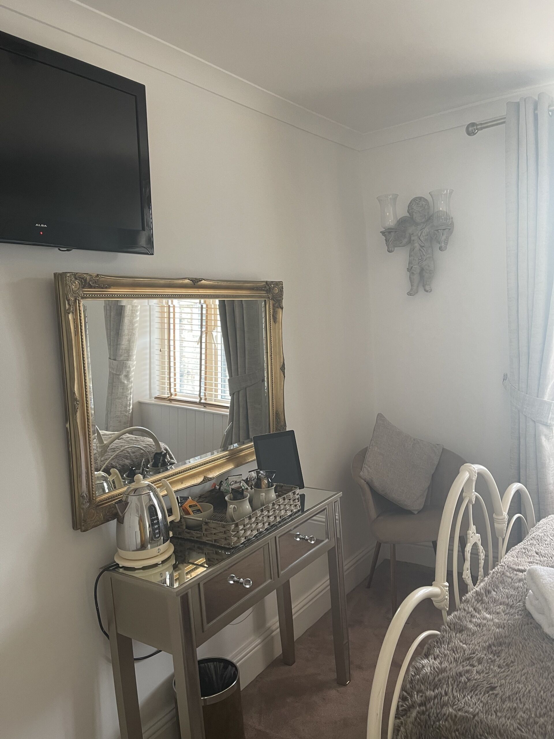 A bedroom with a bed, a bedside table with a kettle, a mirror, a TV, and a window with curtains.