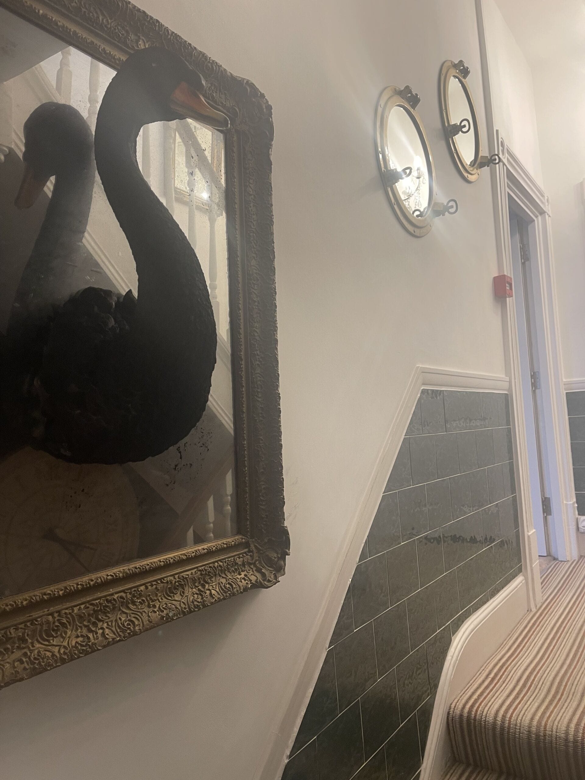 Una escultura de cisne en un espejo, una escalera con baldosas oscuras y una puerta con un botón de emergencia rojo.