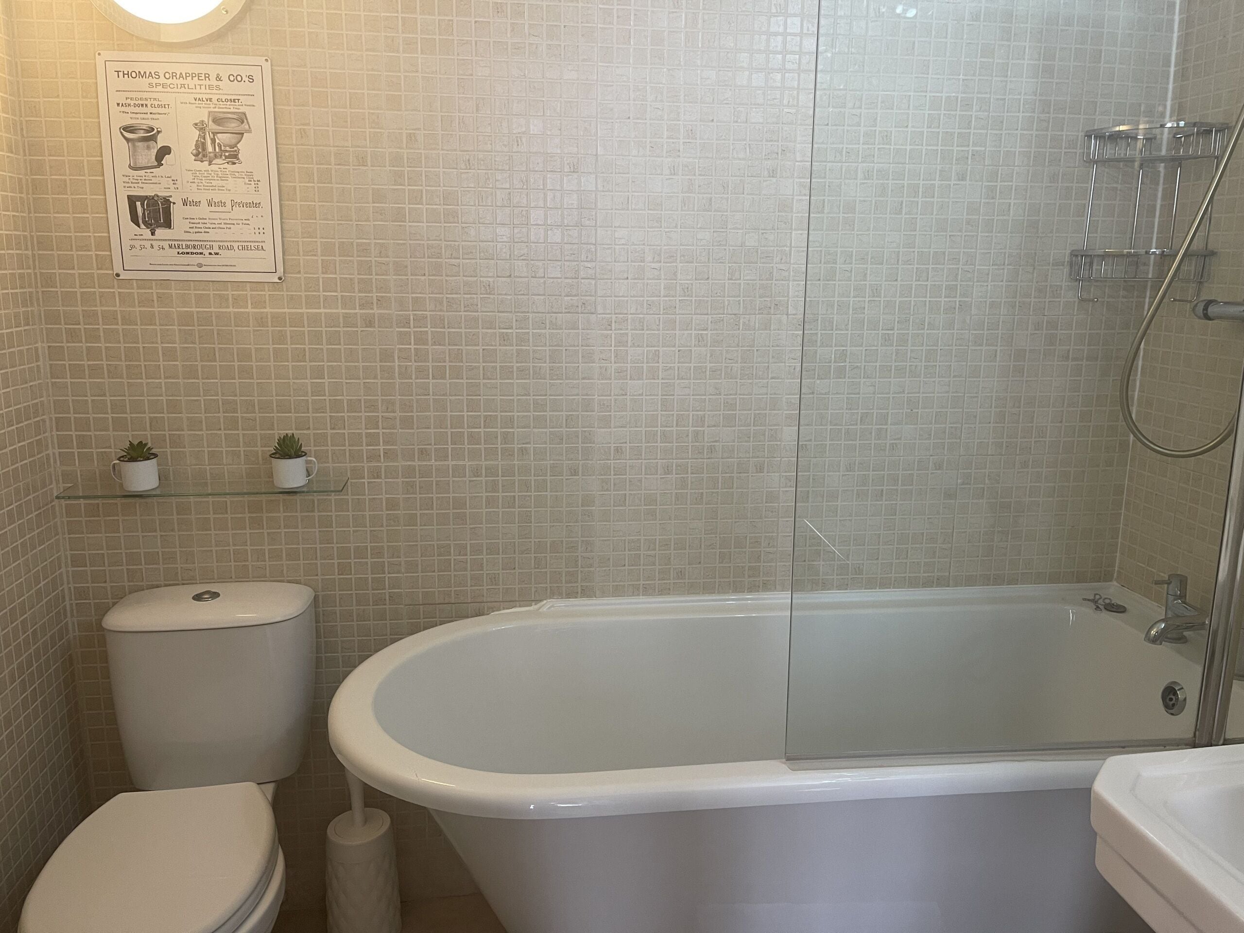 Ein Badezimmer mit einer weißen Badewanne, Toilette und Dusche. Auf einem Regal über der Toilette stehen zwei kleine Topfpflanzen.