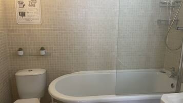 Ein Badezimmer mit einer weißen Badewanne, Toilette und Dusche. Auf einem Regal über der Toilette stehen zwei kleine Topfpflanzen.