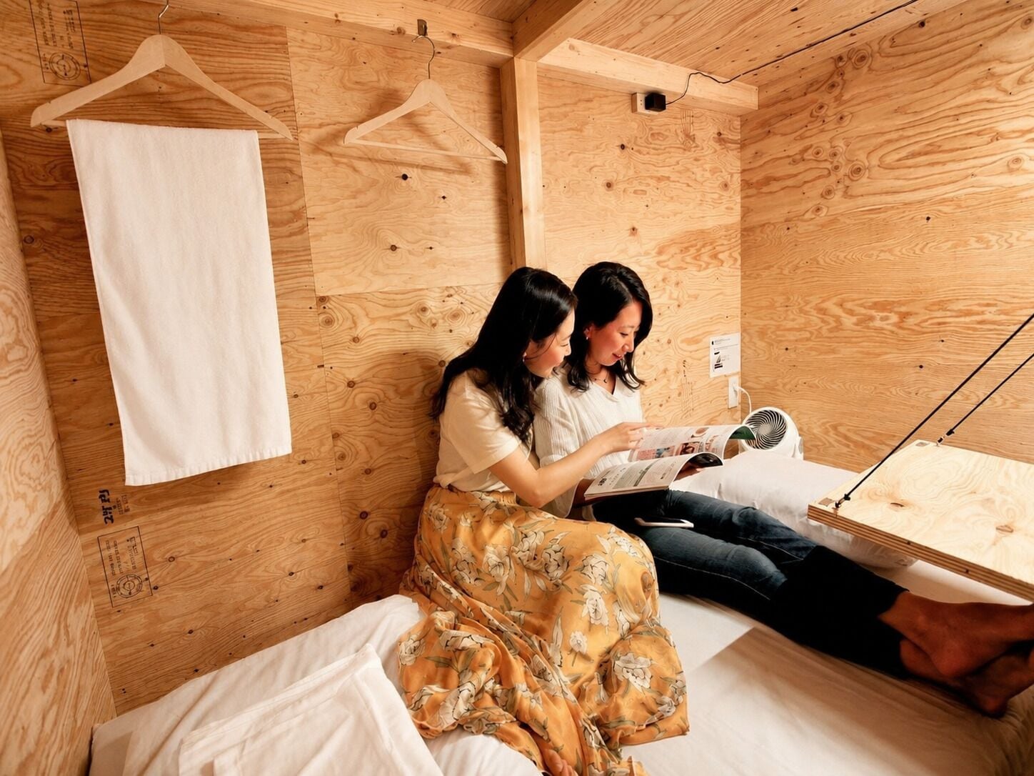A wooden room with a bed, two people reading, and a fan.