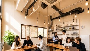 Un grupo de personas en un café moderno con mesas de madera, luces colgantes y una zona de cocina al fondo.