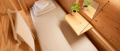 A compact, wooden-furnished room with a bed, a small bedside table with a plant, a mirror, and a wall-mounted fan.