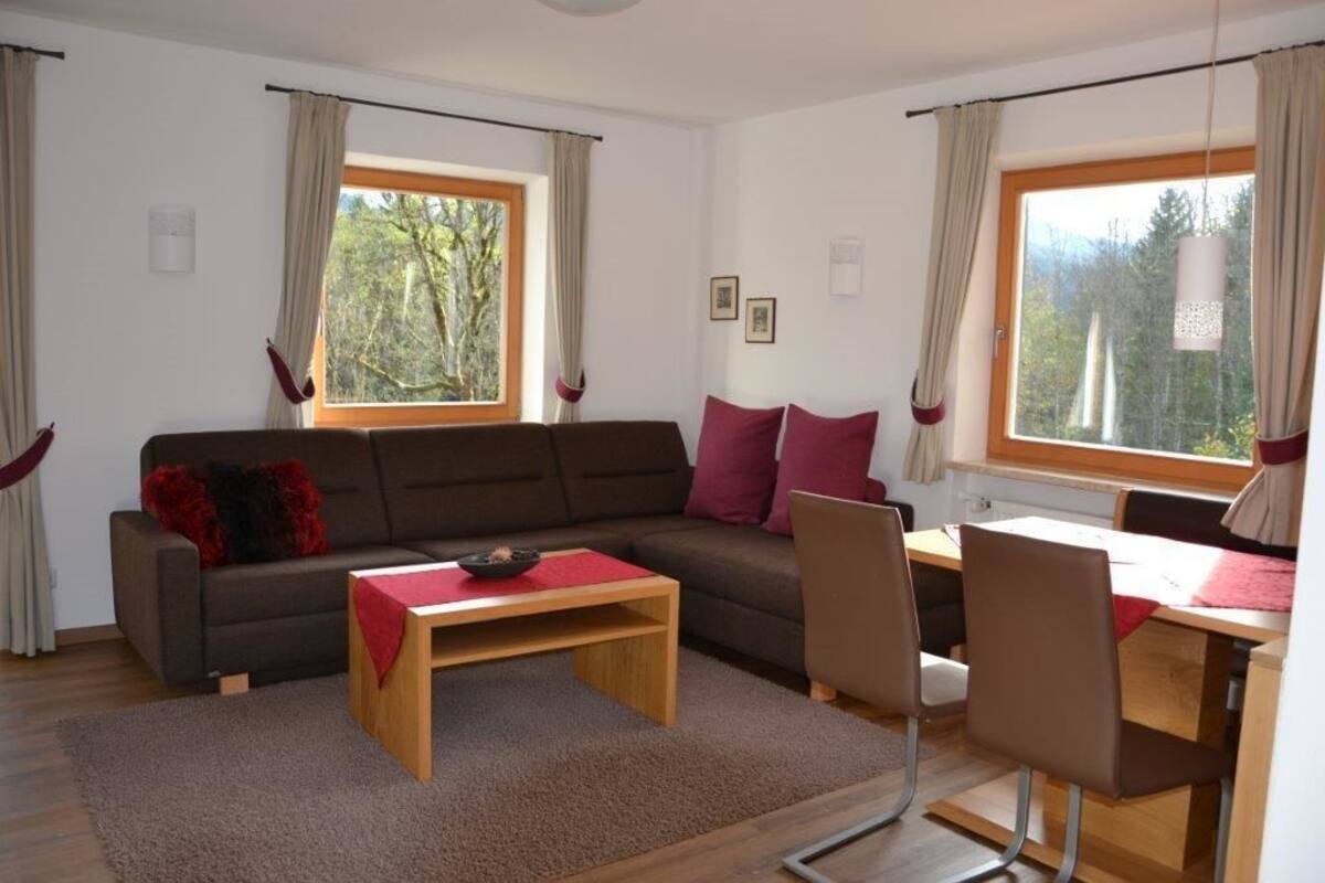 Uma sala de estar moderna com sofá, mesa de centro e área de jantar com cadeiras e mesa. Há duas janelas com cortinas, e o ambiente tem uma paleta de cores neutras.