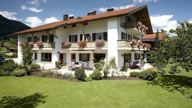 Um edifício de dois andares com varandas, um gramado bem cuidado e montanhas ao fundo.
