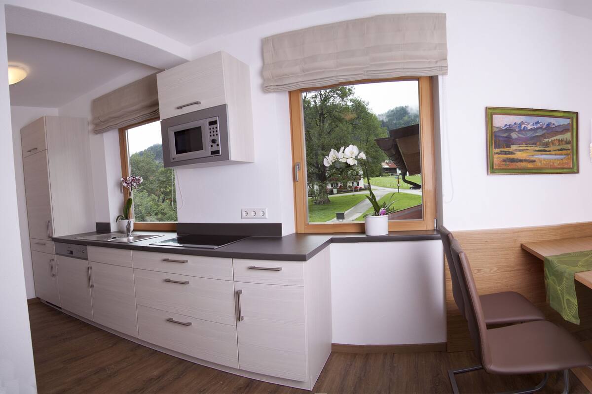 Uma cozinha moderna com micro-ondas embutido, janela com vista para o verde, área de refeições com banco e mesa com toalha de mesa verde.