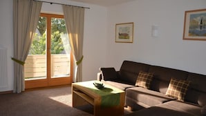 Uma sala de estar com um sofá marrom, uma mesa de centro de madeira e vista para uma área externa através de uma porta de vidro.