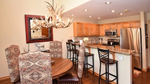 A modern kitchen with wooden cabinets, stainless steel appliances, a central island, and a dining area with patterned chairs and a wooden table.