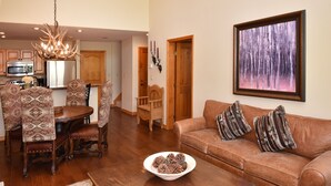 A spacious living room with a brown leather sofa, patterned chairs, a wooden coffee table, and a painting of a forest on the wall.