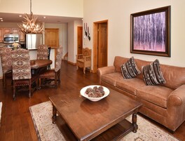 A spacious living room with a brown leather sofa, patterned chairs, a wooden coffee table, and a painting of a forest on the wall.