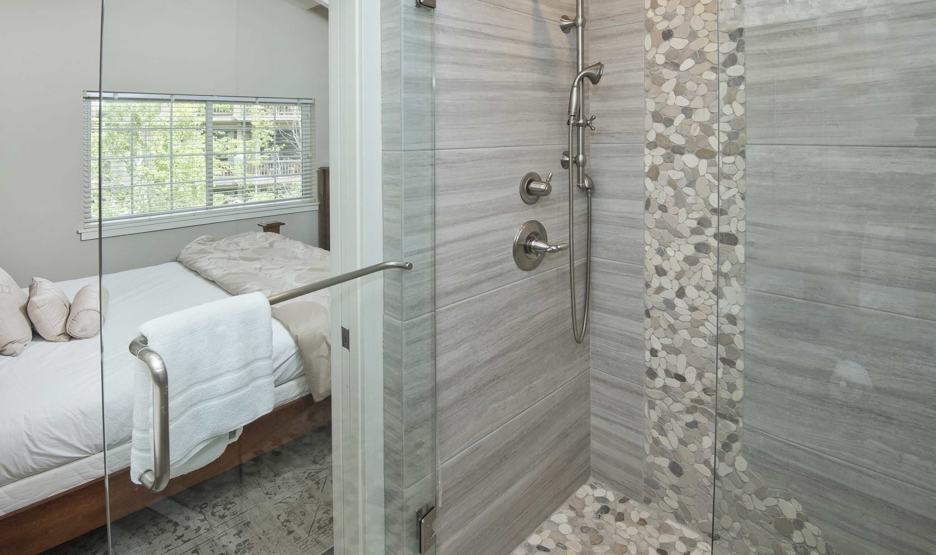A modern bathroom with a glass shower enclosure, a bed with a white towel, and a window with blinds.