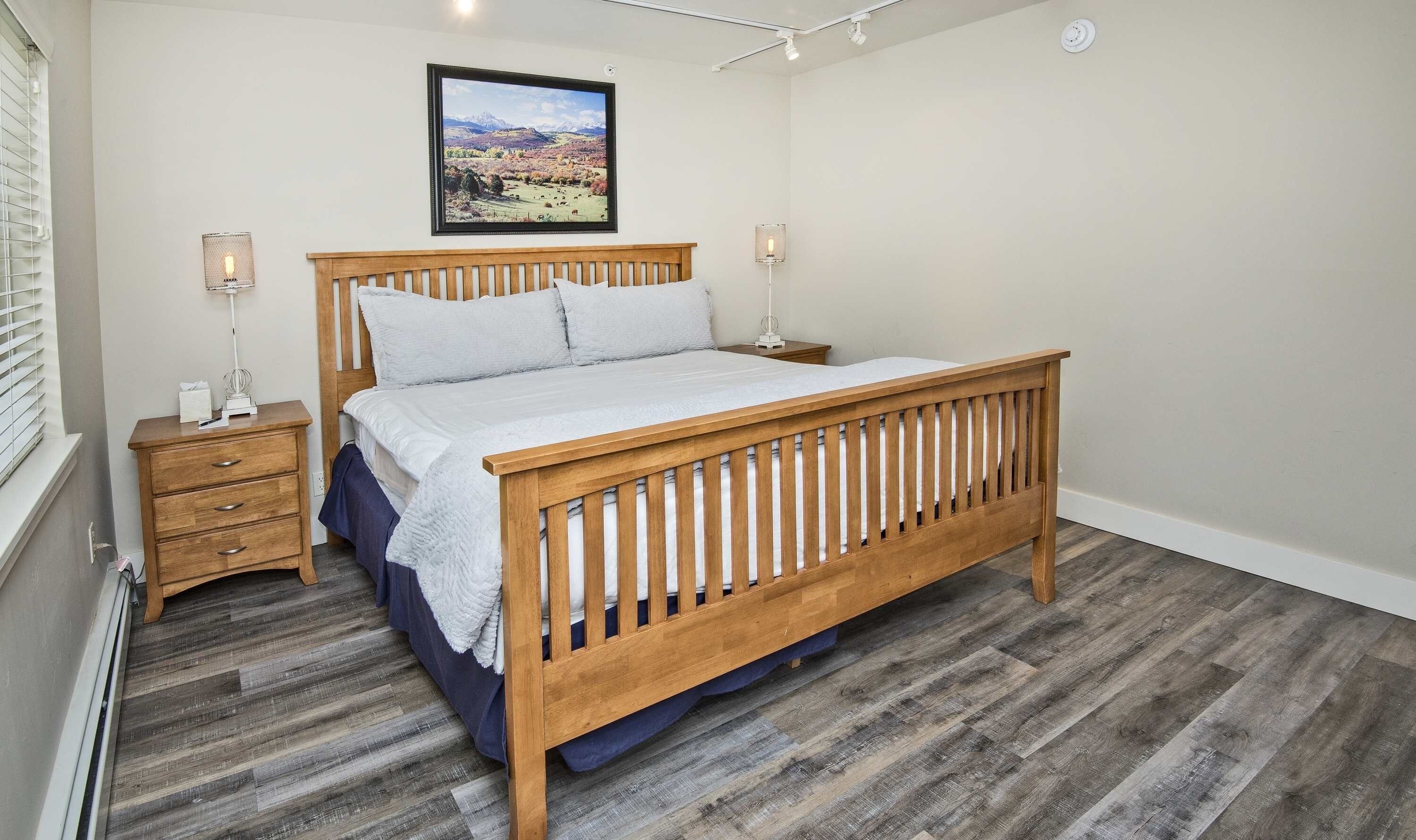 Un dormitorio con cama de madera, mesita de noche, lámpara de noche y un cuadro de paisaje en la pared.