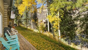 A wooden deck with chairs, a stone fireplace, and a view of a forested area with autumn foliage. - Timbercreek #8b 2 Bedroom Home by RedAwning (Vail)