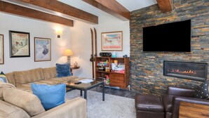 A cozy living room with a stone fireplace, a flat-screen TV mounted on the wall, and a bookshelf filled with books and decorative items. - Vantage Point 112 2 Bedroom Condo by RedAwning (Vail)