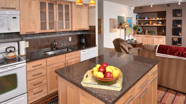 Una cocina con armarios de madera, encimera de granito, un bol de fruta y una cafetera.