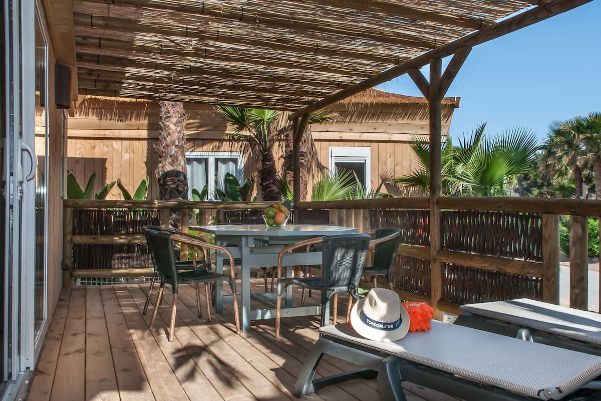 An outdoor patio with a wooden deck, a table with chairs, a hat on a lounge chair, and a thatched roof structure.