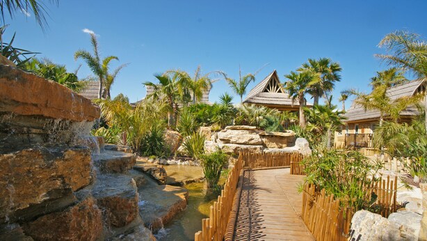 A wooden boardwalk leading to a thatched-roof building surrounded by palm trees and a small water feature. - La Toison d'Or Resort & Spa (Ramatuelle)