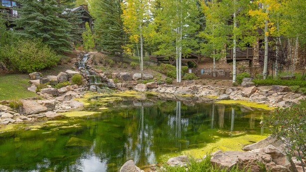 Un étang bordé de rochers et surmonté d’un pont en bois, entouré d’arbres et d’un bâtiment doté d’une terrasse.