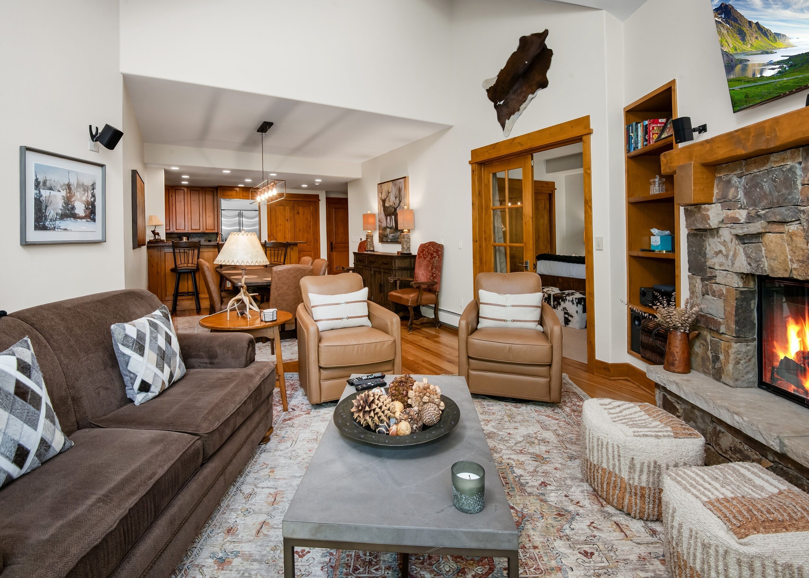 A cozy living room with a fireplace, leather chairs, a coffee table with decorative items, and a kitchen in the background.