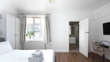 A bedroom with a bed, a desk, a chair, a window with curtains, and a door leading to a bathroom.