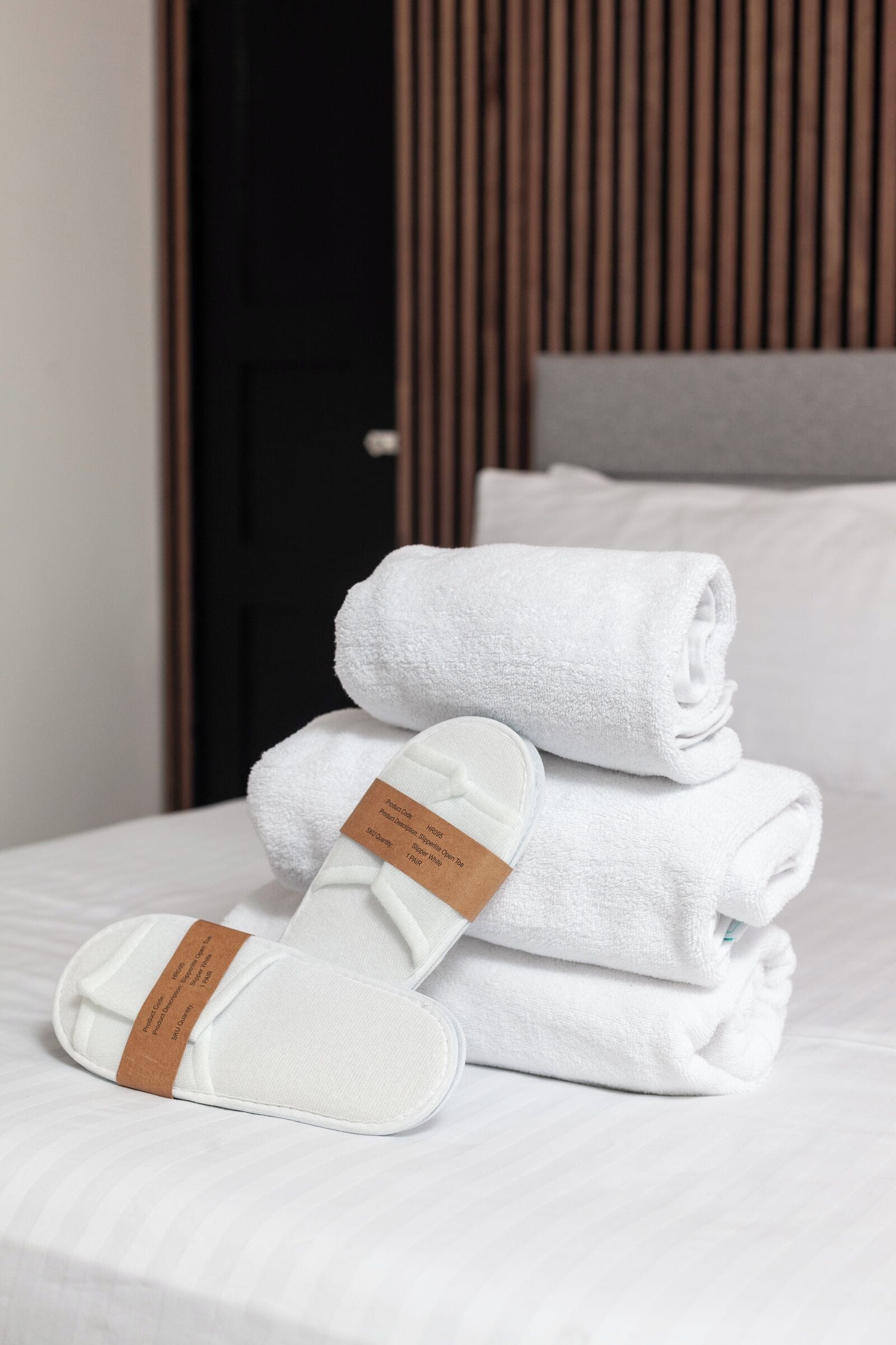 A neatly made bed with white towels and slippers, a wooden headboard, and a white pillow.