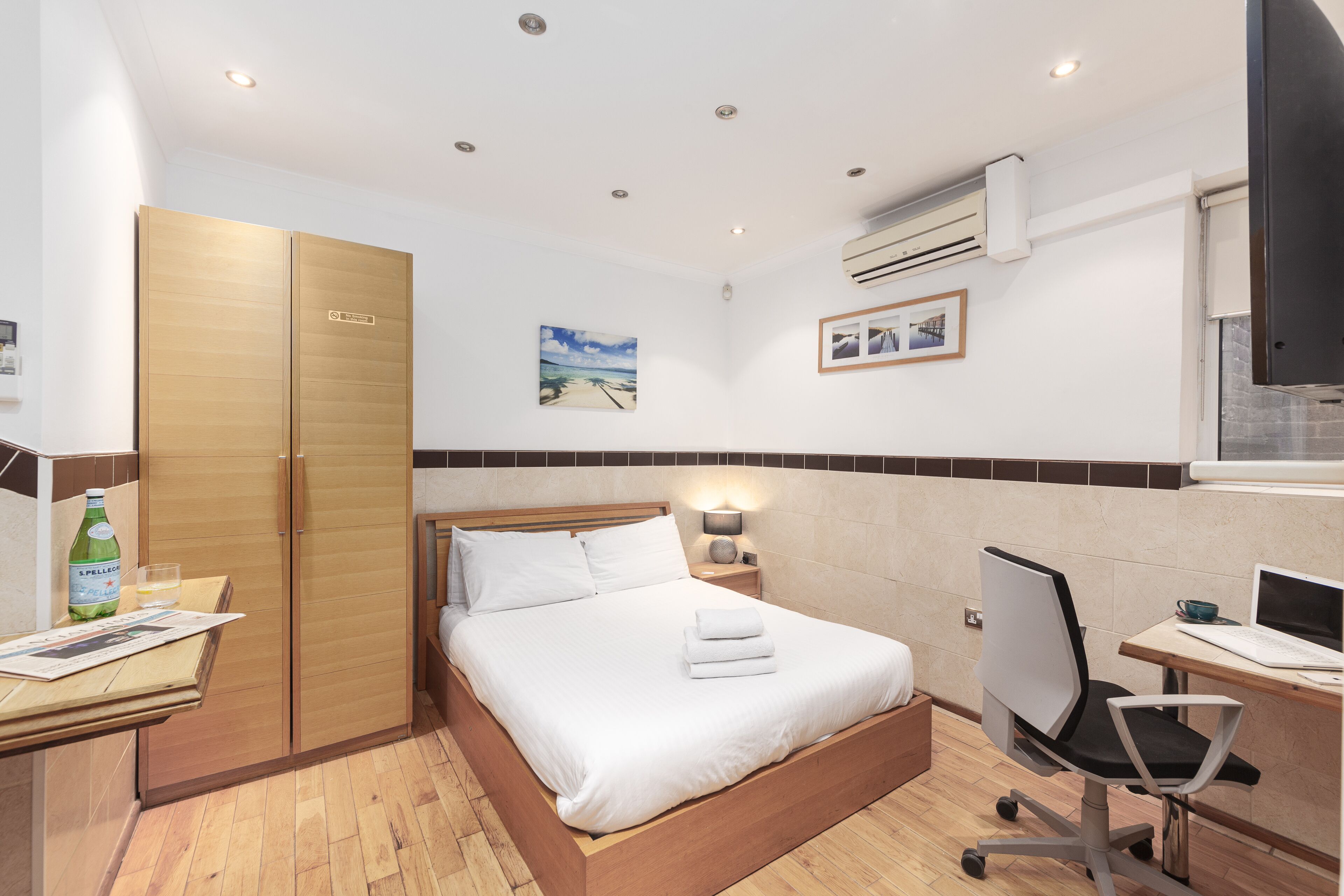 Ein Hotelzimmer mit Bett, Schreibtisch mit Laptop, Stuhl, Kleiderschrank, Wand-Fernseher und einem Bild von einem Strand.