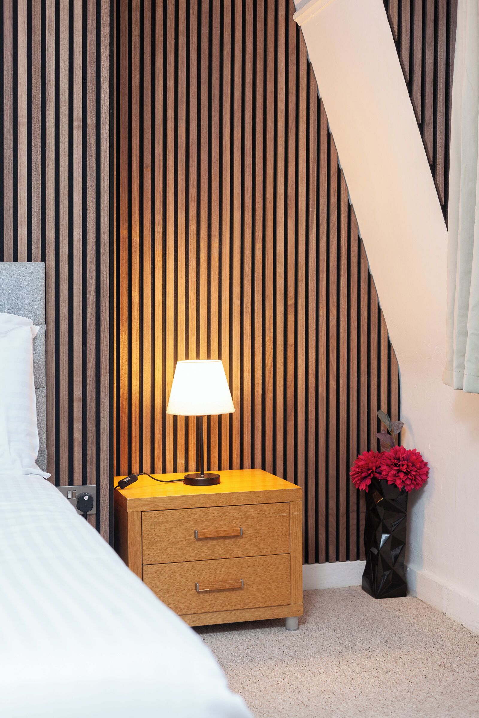 A bedroom with a wooden slat wall, a bedside table with a lamp, a bed with white bedding, and a vase with red flowers.