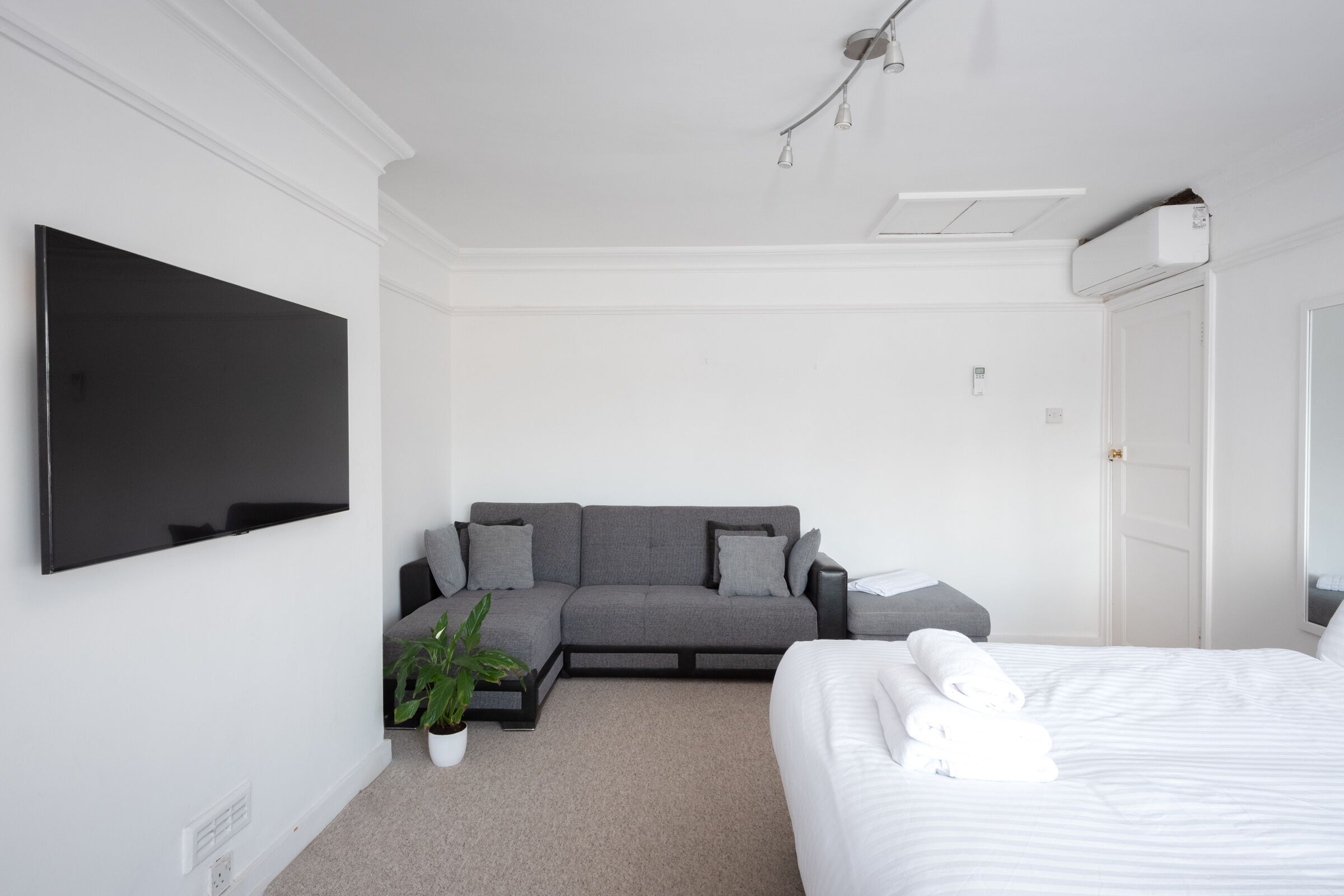 Habitación de hotel moderna con televisión de pantalla plana, sofá gris, planta en maceta y una cama bien hecha con toallas.