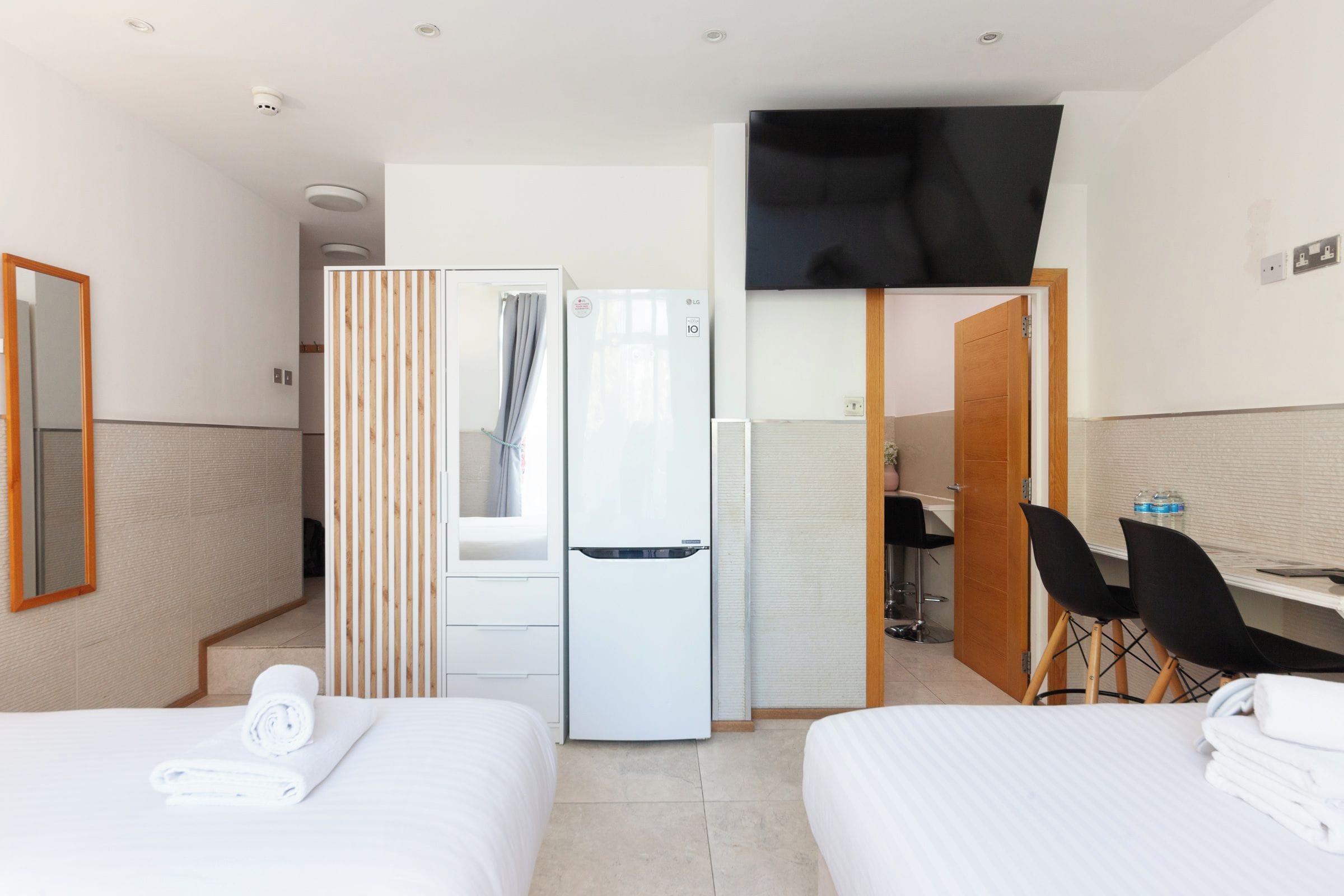 A hotel room with two beds, a refrigerator, a flat-screen TV, a wooden partition, a desk with a chair, and a mirror.