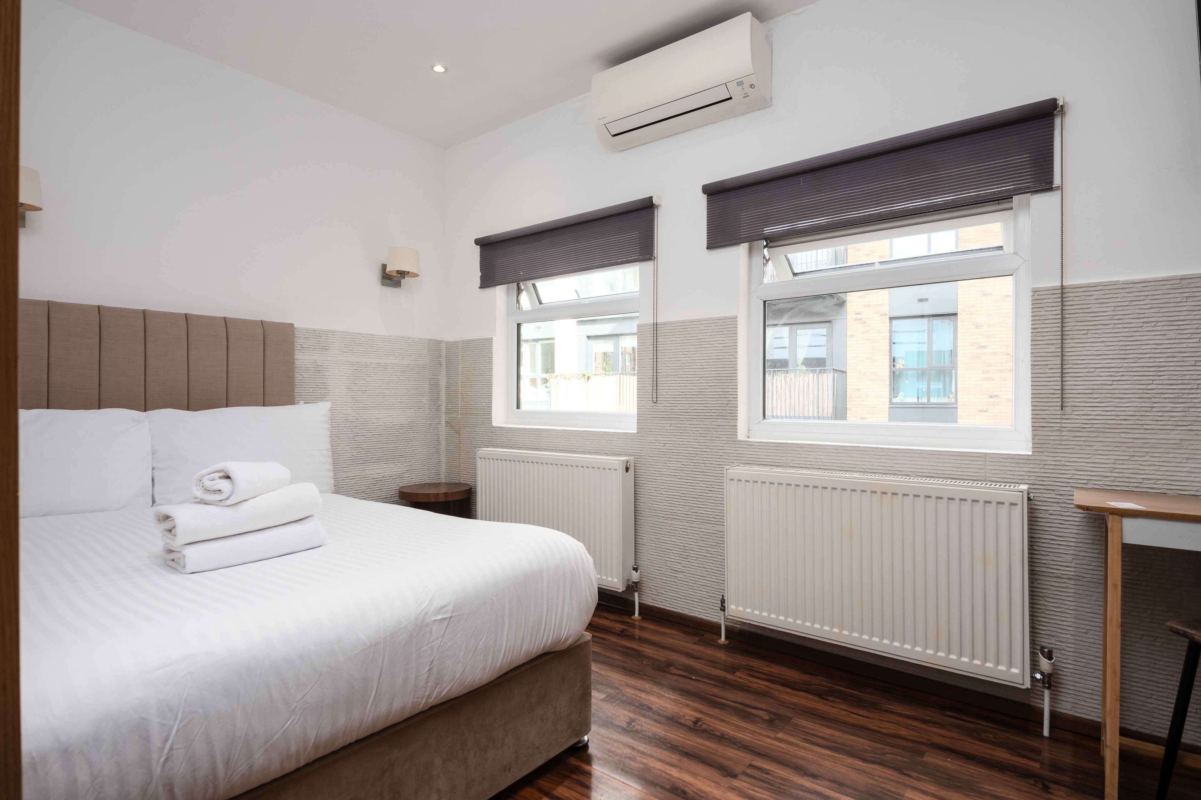 Ein Schlafzimmer mit einem Bett, zwei Fenstern mit Jalousien, einer Klimaanlage und einem Heizkörper.