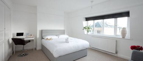 Ein Schlafzimmer mit einem Bett, einem Schreibtisch mit einem Laptop, einem Stuhl, einem Heizkörper und einem Fenster mit Blick auf Gebäude.