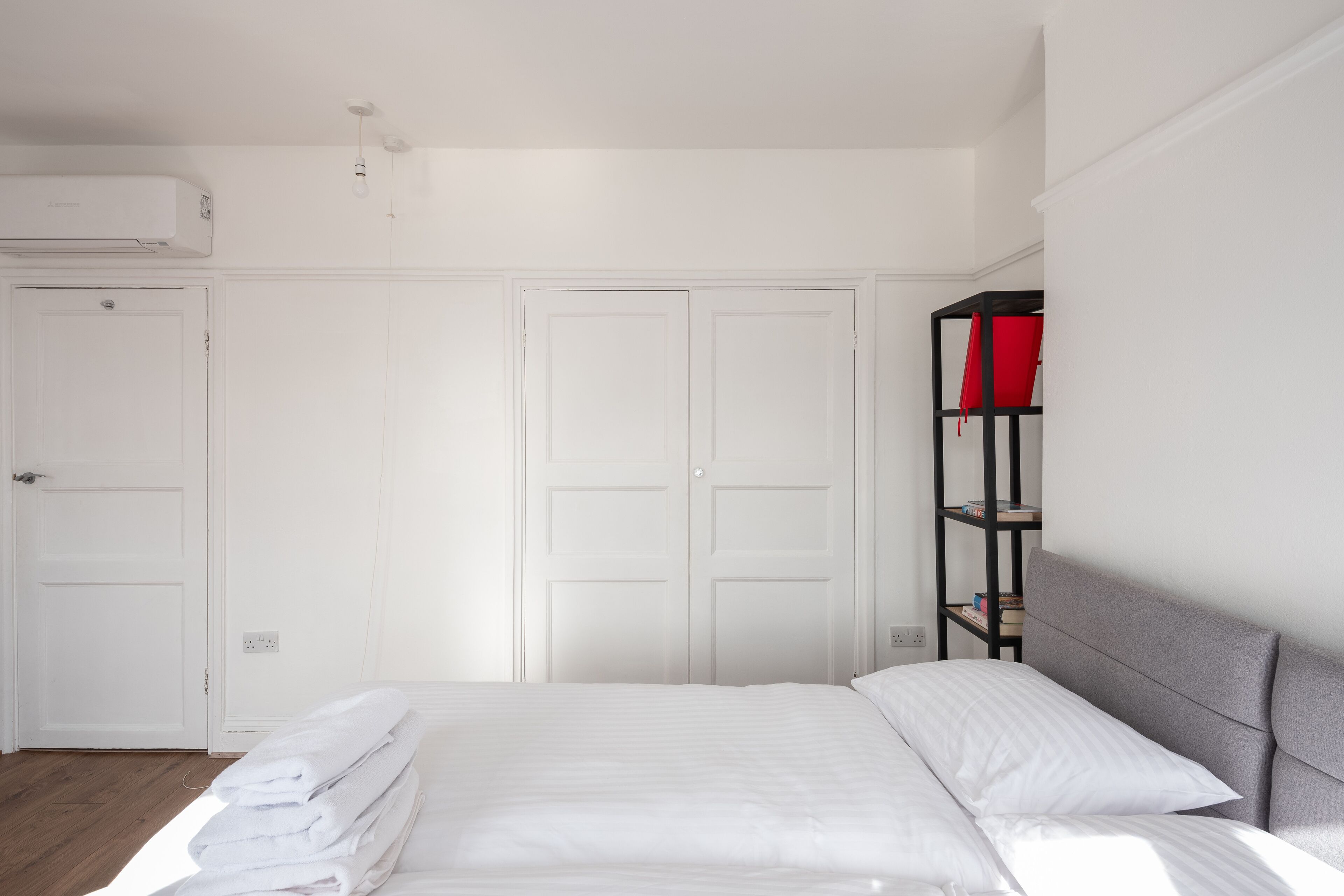 Un dormitorio con una cama bien hecha, paredes blancas, un armario blanco y una estantería negra con cojines rojos.