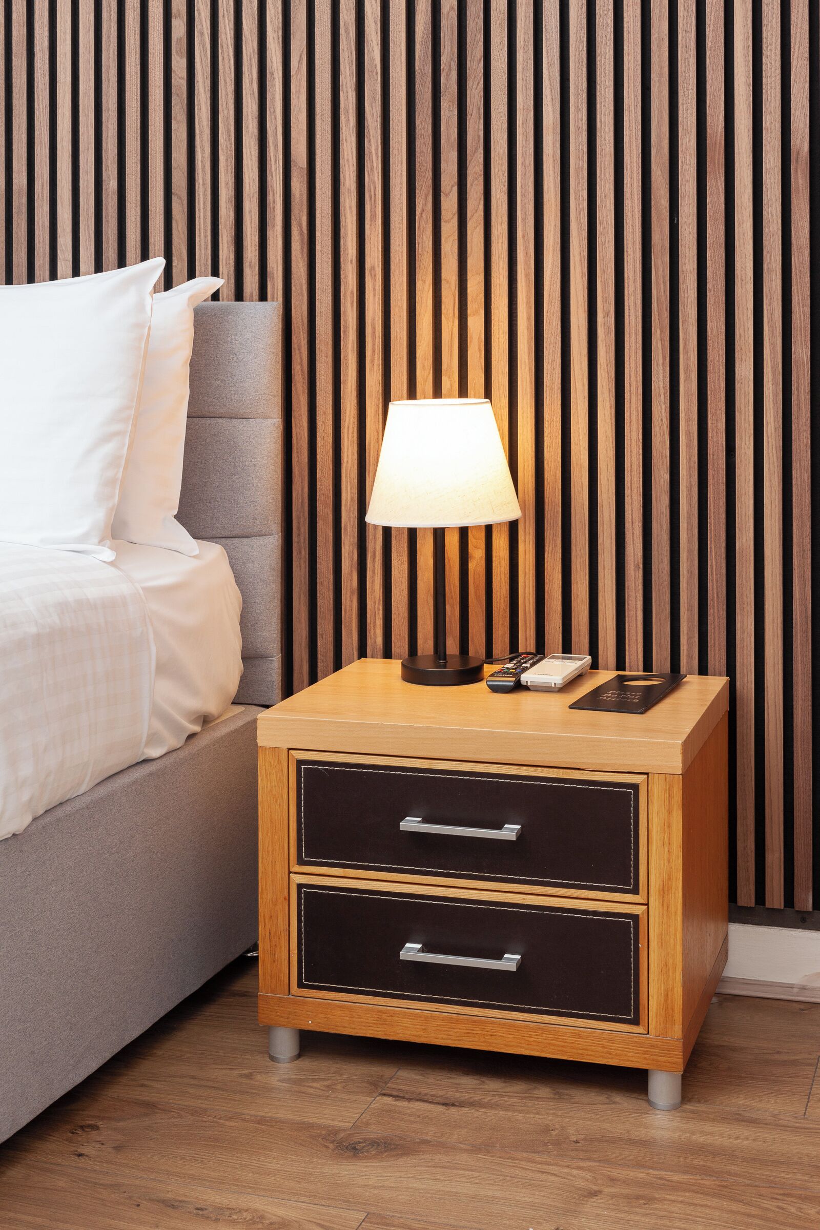 A neatly made bed with white linens, a bedside lamp, a small bedside table with drawers, and a wooden slatted wall behind.